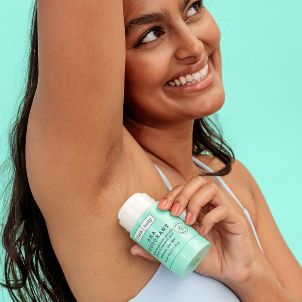 A smiling woman with her arm raised holding a frank body AHA Deodorant stick.