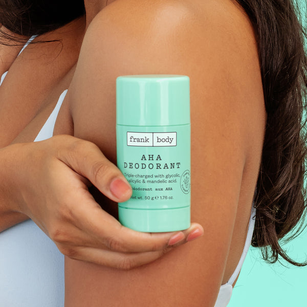 A woman holding a frank body AHA Deodorant stick against her upper arm.