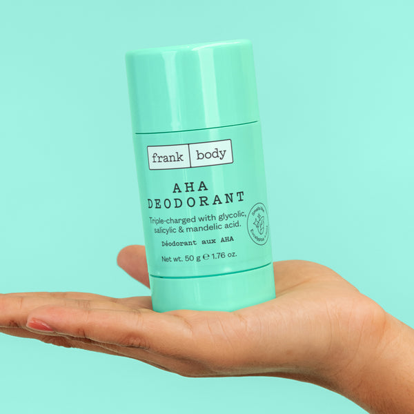 A hand holding a frank body AHA Deodorant stick on a mint background.