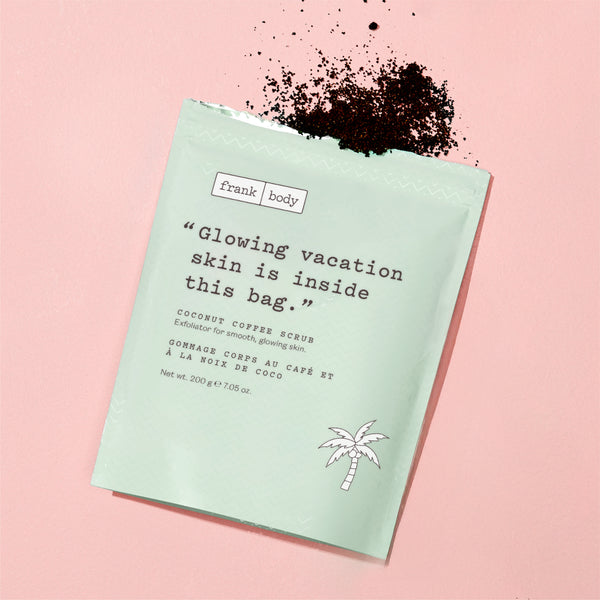A mint green stand-up pouch of Frank Body Coconut Coffee Scrub lies on a soft pink background with dark coffee powder scattered on top. The package features black typewriter-style text and a small palm tree illustration.