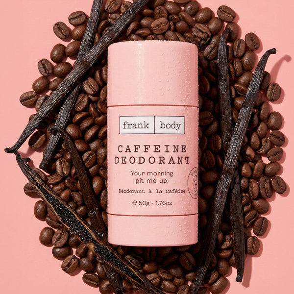A frank body Caffeine Deodorant stick surrounded by coffee beans and vanilla pods on a pink background.