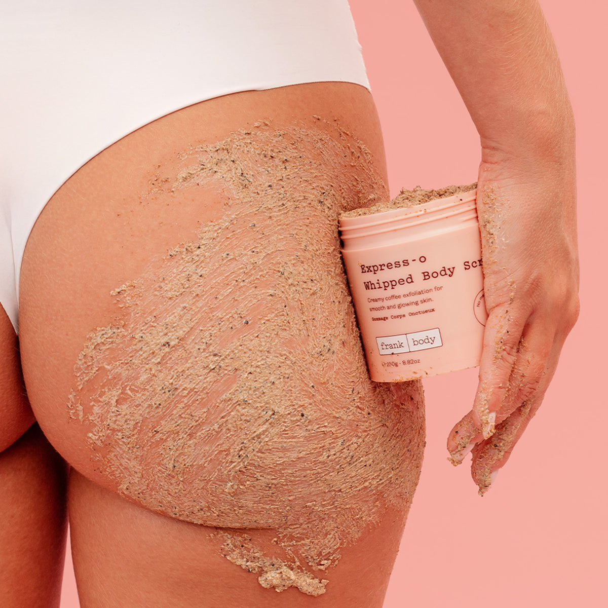 A woman in white underwear with frank body Express-o Whipped Body Scrub across her hip, holding the tub.